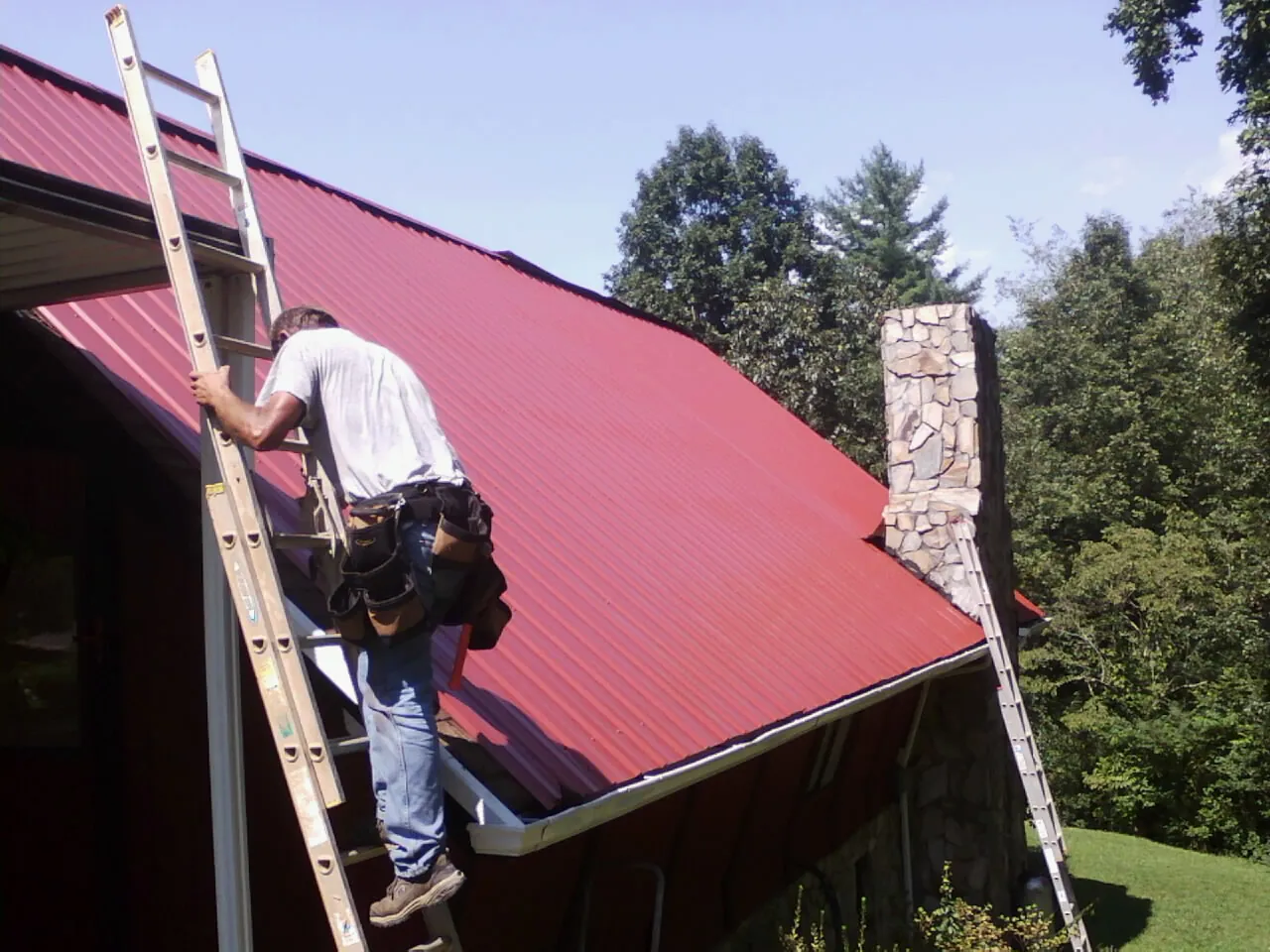 Roofer on a ladder beside a red metal roof and stone chimney — Soffit & Fascia Repair by Roofing Brothers in Poynette